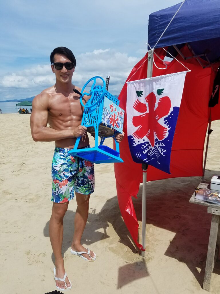 海辺でかき氷機を手にしたマッチョと「氷」ののぼり