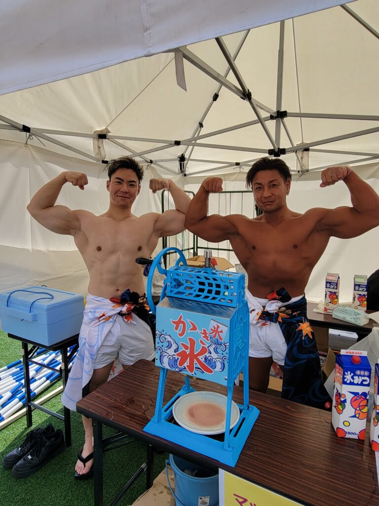 青いかき氷機の横で2人のメンバーがポーズをとっている様子
